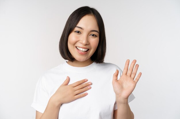 Porträt eines schönen koreanischen Mädchens, das die Hand hebt, stellt sich vor und legt die Hand auf den Herzgruß, der über weißem Hintergrund steht