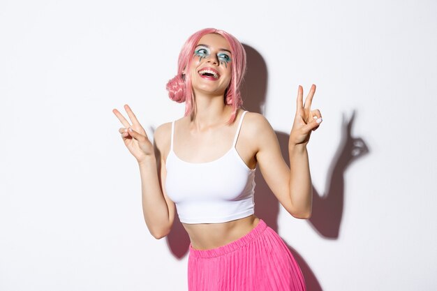 Porträt eines schönen lächelnden Mädchens in rosa Perücke, das kawaii Friedenszeichen zeigt und lacht, Outfit für Party trägt und auf weißem Hintergrund steht