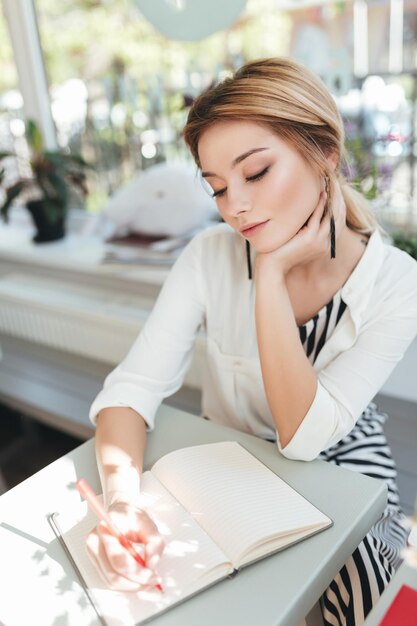 Porträt eines schönen Mädchens, das im Café sitzt und verträumt ihre Augen mit einem Notizbuch auf dem Tisch schließt. Hübsches Mädchen mit blonden Haaren, das eine Notiz am Tisch im Restaurant schreibt