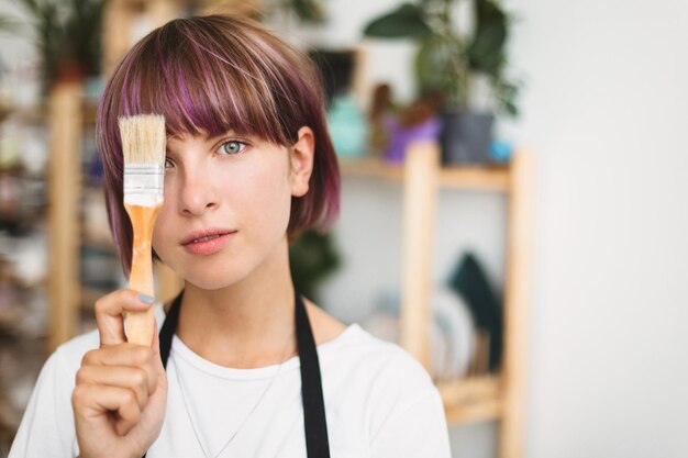 Porträt eines schönen Mädchens mit bunten Haaren im weißen T-Shirt, das ihr Auge mit einem Pinsel bedeckt, der verträumt in die Kamera in das Töpferstudio blickt