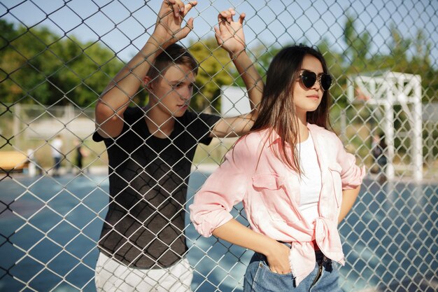 Porträt eines schönen Mädchens mit dunklem Haar und Sonnenbrille, das neben einem Maschendrahtzaun und einem coolen Jungen im Hintergrund auf einem Basketballplatz steht