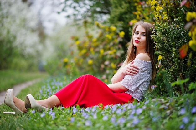 Porträt eines schönen Mädchens mit roten Lippen im Frühlingsblütengarten auf Gras mit Blumen auf rotem Kleid und weißer Bluse