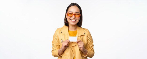 Porträt eines schönen modernen asiatischen Mädchens mit Sonnenbrille, das glücklich lächelt und eine Kreditkarte zeigt, die über weißem Hintergrundkopienraum steht