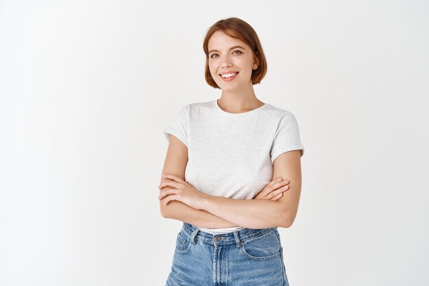 Porträt eines schönen natürlichen weiblichen Modells mit kurzen Haaren, T-Shirt mit Jeans, verschränkten Armen auf der Brust und einem Lächeln mit glücklichen Emotionen, weiße Wand