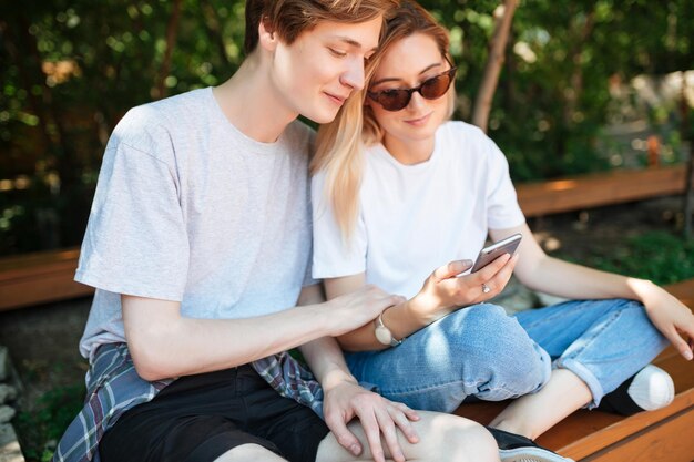Porträt eines schönen Paares, das auf einer Bank im Park sitzt und ein Mobiltelefon benutzt Nahaufnahme eines jungen Mannes und einer hübschen Dame mit blonden Haaren, die das Mobiltelefon in der Hand halten, während sie Zeit miteinander verbringen