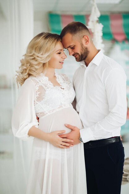 Porträt eines schönen Paares von Mutter und Vater, die den Bauch umarmen und sich gegenseitig von Angesicht zu Angesicht anschauen Schwangere blonde Frau in Weiß lächelt ihrem gutaussehenden Mann zu, der sich in einem stilvollen Interieur ausgibt