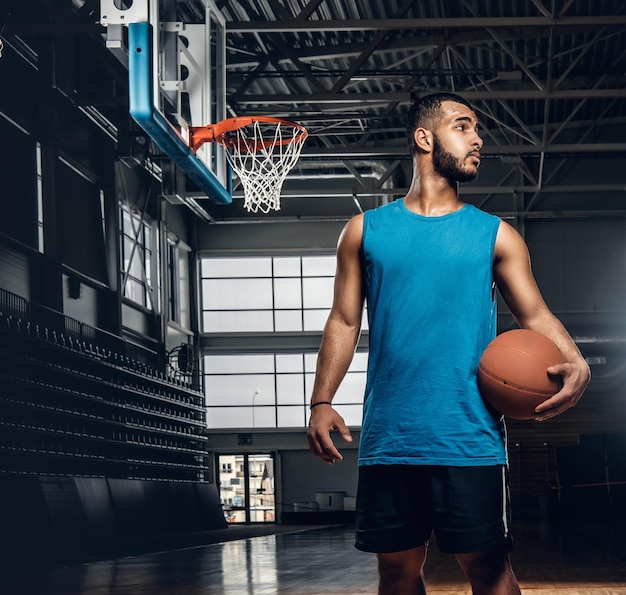 Kostenloses Foto porträt eines schwarzen basketballspielers hält einen ball über einem reifen in einer basketballhalle.