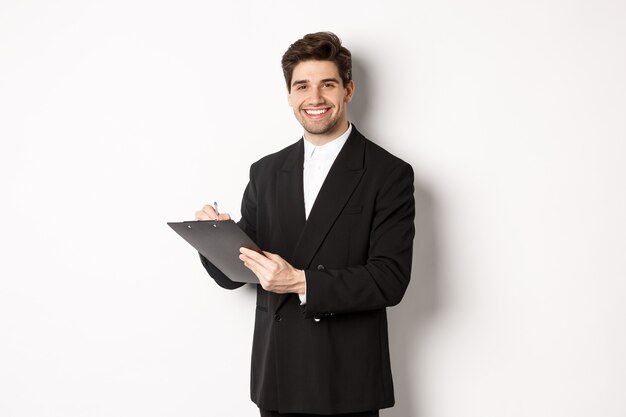Porträt eines selbstbewussten Geschäftsmannes im schwarzen Anzug, der Dokumente unterschreibt und lächelt und vor weißem Hintergrund glücklich steht.