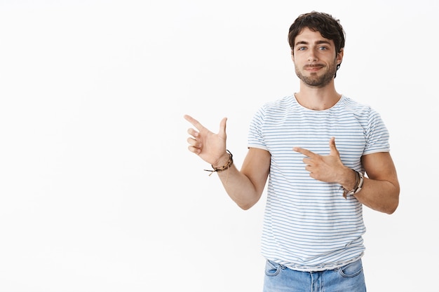 Porträt eines selbstbewussten, gut aussehenden, gebräunten jungen kaukasischen Mannes mit Borsten, blauen Augen in gestreiftem T-Shirt, das mit Fingerpistolen zeigt, lächelt zufrieden und posiert zufrieden gegen graue Wand