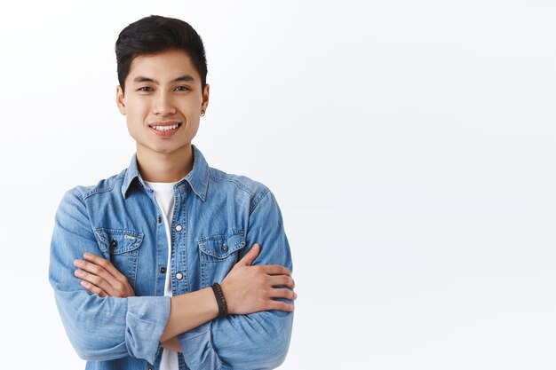 Porträt eines selbstbewussten jungen Hipster-Männchens, asiatischer Kerl in Jeansjacke, der mit Freunden auf dem Campus spricht, überquert die lässige Brustpose, lächelt zufrieden, empfiehlt Sprachkurse, weiße Wand
