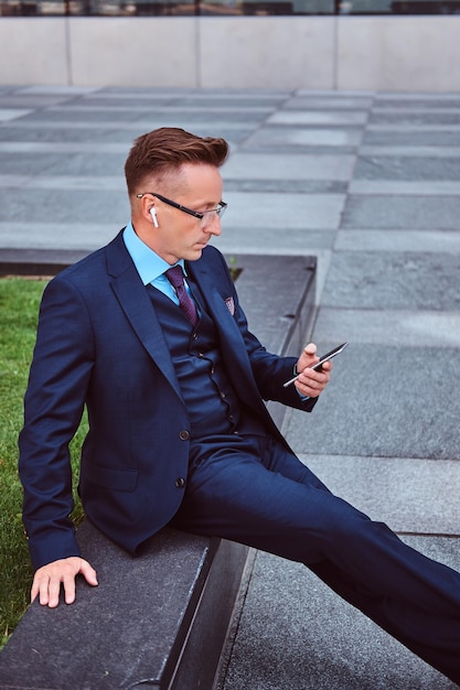 Porträt eines selbstbewussten, stilvollen Geschäftsmannes in einem eleganten Anzug, der ein Smartphone benutzt, während er im Freien vor einem Wolkenkratzerhintergrund sitzt.