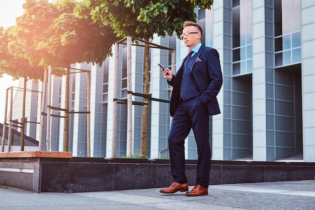 Porträt eines selbstbewussten, stilvollen Geschäftsmannes in einem eleganten Anzug hält ein Smartphone und schaut weg, während er im Freien vor Wolkenkratzerhintergrund steht.