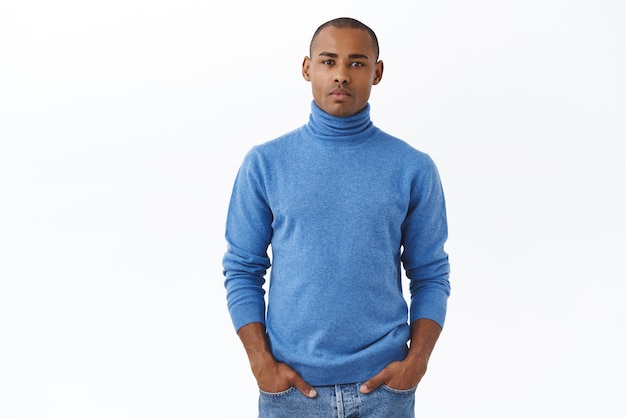Porträt eines seriös aussehenden, selbstbewussten jungen Mannes in blauem Rollkragenpullover mit Borstenhändchen in Jeanstaschen und kameraschauendem, entschlossenem Hören auf den denkenden weißen Hintergrund des Chefs