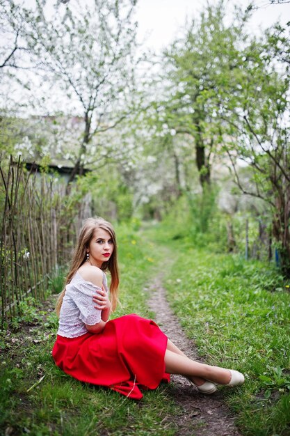 Porträt eines sitzenden schönen Mädchens mit roten Lippen im Frühlingsblütengarten auf grünem Gras auf rotem Kleid und weißer Bluse