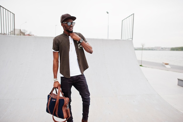 Porträt eines sitzenden stilvollen afroamerikanischen Mannes mit Sonnenbrille und Mütze mit Handtasche im Freien gegen den Skatepark Straßenmode schwarzer Mann