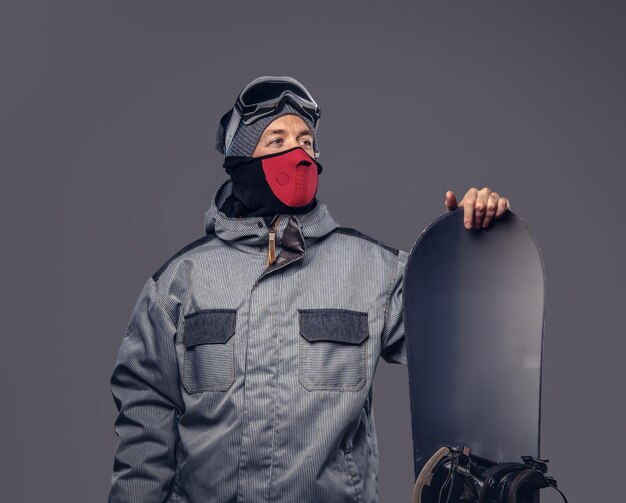 Porträt eines Snowboarders in voller Schutzausrüstung für extremes Snowboarden, der mit einem Snowboard in einem Studio posiert. Isoliert auf grauem Hintergrund.