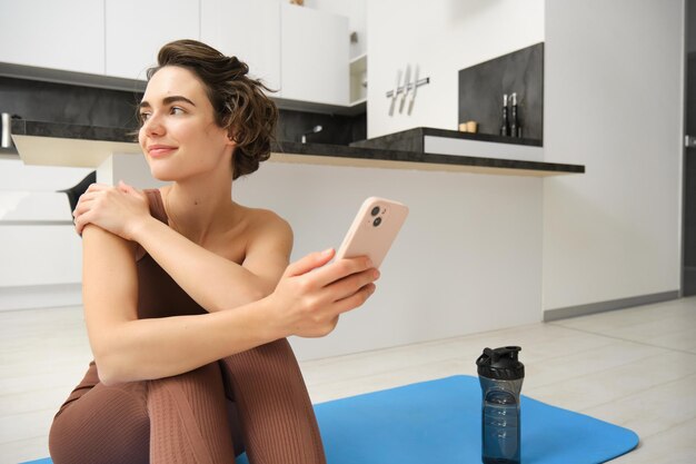 Porträt eines sportlichen Mädchens mit Mobiltelefon, das während des Trainings zu Hause auf einer Yogamatte sitzt und sein Smartphone nutzt