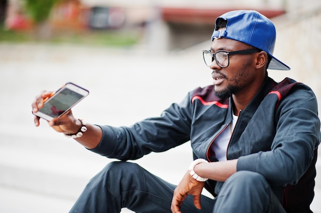 Porträt eines stilvollen afroamerikanischen Mannes mit Sportswear-Mütze und Brille, der mit dem Telefon zur Hand auf der Treppe sitzt Schwarze Männer modellieren Straßenmode