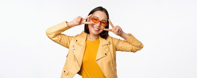 Porträt eines stilvollen asiatischen modernen Mädchens mit Sonnenbrille und gelber Jacke, das eine friedliche Vsign-Geste zeigt, die über einem glücklich lächelnden Gesicht des weißen Hintergrunds steht