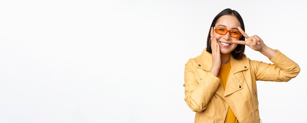 Porträt eines stilvollen asiatischen modernen Mädchens mit Sonnenbrille und gelber Jacke, das eine friedliche Vsign-Geste zeigt, die über einem glücklich lächelnden Gesicht des weißen Hintergrunds steht