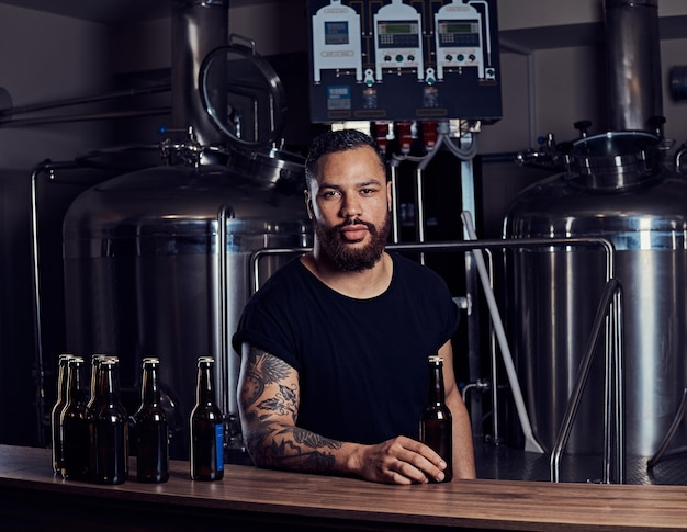 Porträt eines stilvollen, bärtigen, dunkelhäutigen Mannes mit einer Tätowierung auf der Hand, der hinter der Theke in einer Brauerei steht.