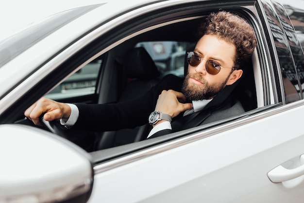 Porträt eines stilvollen bärtigen Mannes mit Sonnenbrille hinter dem Steuer eines weißen Autos, eine teure Uhr zur Hand