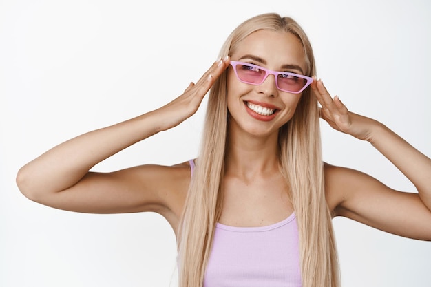 Kostenloses Foto porträt eines stilvollen blonden mädchens mit rosa sonnenbrille, das glücklich lächelt und mit sorglosem gesichtsausdruck auf weißem hintergrund zur seite schaut