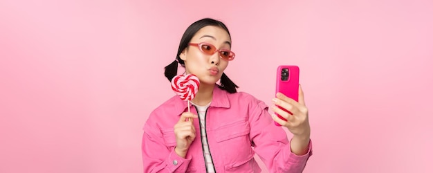 Porträt eines stilvollen, glücklichen asiatischen Mädchens, das ein Selfie mit Süßigkeiten aus Lolipop macht und lächelt, während es fotografiert