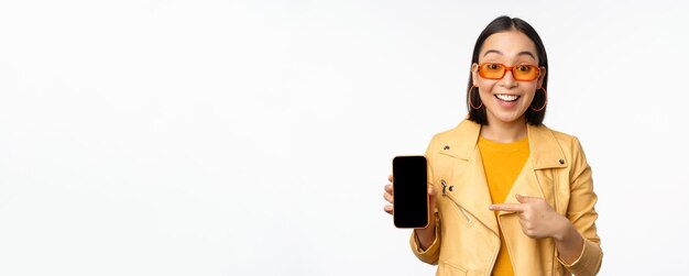 Porträt eines stilvollen koreanischen Mädchens mit Sonnenbrille, das lächelnd mit dem Finger auf den Smartphone-Bildschirm zeigt, auf dem die Handyanwendung vor weißem Hintergrund zu sehen ist