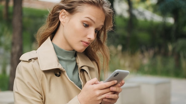 Porträt eines stilvollen Mädchens, das überrascht aussieht, als es auf Nachrichten auf dem Smartphone im Freien antwortet Studentin, die sich im Park ausruht