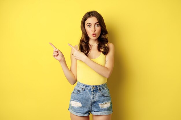 Porträt eines stilvollen Sommermädchens, einer brünetten Frau, die den Weg zeigt, mit dem Finger nach links zeigt, Werbung macht und auf gelbem Hintergrund steht