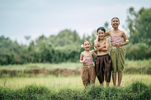 Porträt eines süßen Jungen ohne Hemd mit gekreuzten Armen und zwei hübschen Mädchen in traditioneller thailändischer Kleidung legen eine schöne Blume auf ihr Ohr, die im Reisfeld steht, lachen, Platz kopieren