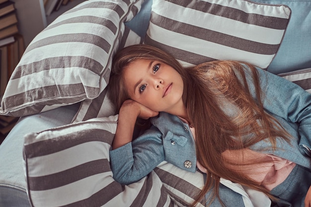 Porträt eines süßen kleinen Mädchens mit langen braunen Haaren und durchdringendem Blick, das auf eine Kamera blickt und allein zu Hause auf einem Sofa liegt.