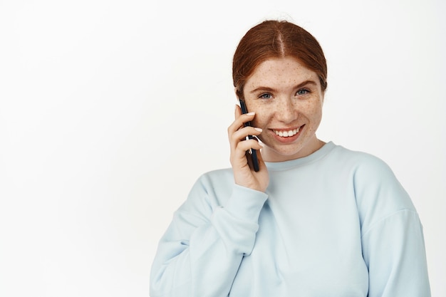 Porträt eines süßen lächelnden rothaarigen Mädchens, das auf dem Handy spricht, mit einem Freund auf dem Handy chatten, Smartphone benutzen, anrufen, auf Weiß stehen
