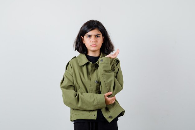 Porträt eines süßen Teenager-Mädchens, das die Handfläche beiseite ausbreitet, während es in der grünen Armeejacke schmollend und verwirrte Vorderansicht schaut