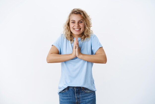 Porträt eines süßen und freundlichen, glücklichen jungen blonden Mädchens mit blauen Augen, das angenehm lächelt, als ob es die Handflächen zusammenhält