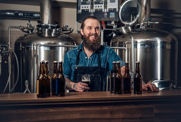 Porträt eines tätowierten, bärtigen Hipster-Herstellers, der eine Mikrobrauerei für Bier präsentiert.