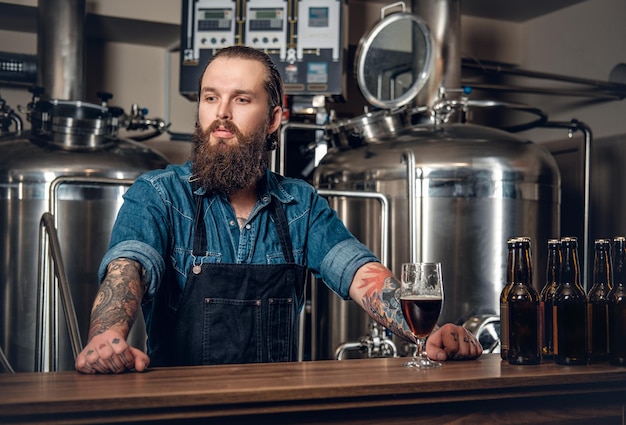 Porträt eines tätowierten, bärtigen Hipster-Herstellers, der eine Mikrobrauerei für Bier präsentiert.