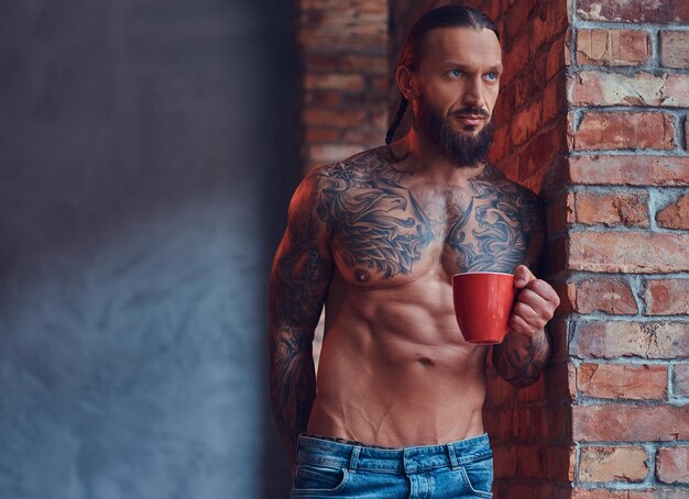 Porträt eines tätowierten, hemdlosen Mannes mit stilvollem Haarschnitt und Bart, trinkt Morgenkaffee und lehnt sich an eine Ziegelwand in einem Raum.