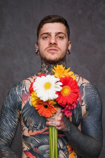 Porträt eines tätowierten Mannes mit dem Durchbohren in den Ohren und in der Nase, die bunte Gerbera in der Hand halten, blüht in der Hand