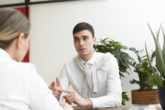 Porträt eines talentierten talentierten jungen männlichen Bewerbers, der einen Termin bei einer Personalvermittlerin hat, während er sich für eine Position als Designer in einem großen Unternehmen bewirbt, mit den Fingern zählt und seine Stärken auflistet