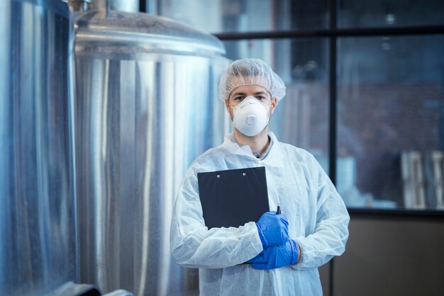 Porträt eines Technologen in weißer Uniform mit Haarnetz und Schutzmaske und Handschuhen, die in der Pharma- oder Lebensmittelfabrik mit verschränkten Armen stehen