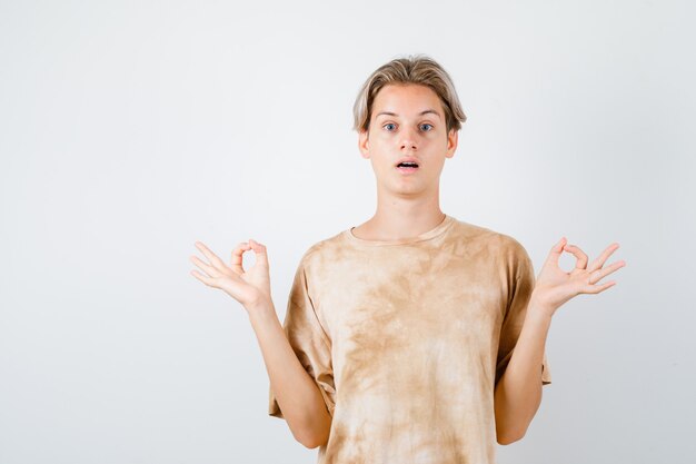 Porträt eines Teenagerjungen, der Mudra-Zeichen im T-Shirt zeigt und verwirrte Vorderansicht schaut