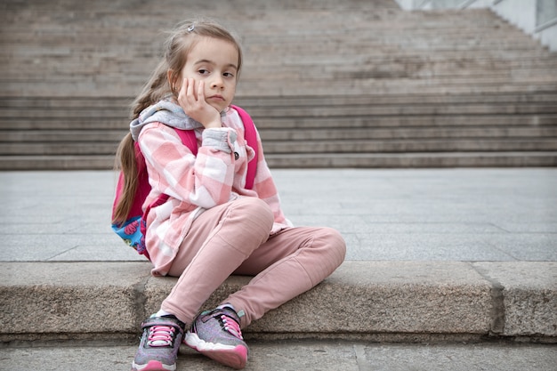Porträt eines traurigen kleinen Mädchens mit einer Aktentasche auf dem Rücken, das auf der Treppe sitzt. Zurück zur Schule.