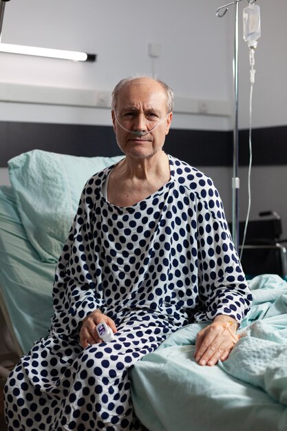 Porträt eines traurigen, unwohlen älteren Mannes, der am Rand des Krankenhausbettes mit angebrachtem iv-Tropf sitzt und mit Hilfe einer Sauerstoffmaske atmet, nach vorne schauend