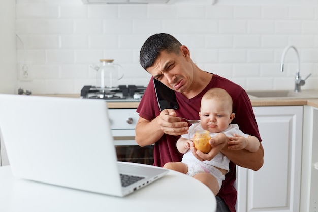 Porträt eines traurigen verärgerten Mannes, der ein kastanienbraunes Casual-T-Shirt trägt, das mit einer kleinen Tochter oder einem Sohn auf Knien sitzt, telefoniert und weint, braucht Arbeit und kümmert sich um das Baby, hat keine Zeit.