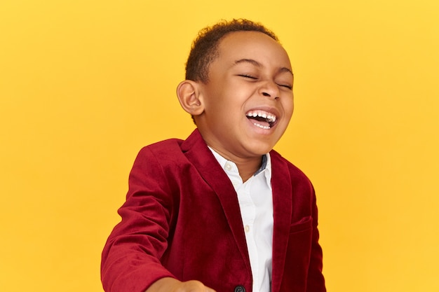 Porträt eines überglücklichen glücklichen afrikanischen kleinen Jungen in der trendigen Jacke, der Kopf zurück wirft, während über Witz laut lacht, Spaß hat