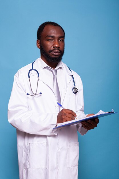 Porträt eines übermütigen Arztes, der in die Kamera blickt und ein Klemmbrett mit Patientenakten hält. Selbstbewusster Sanitäter, der im Laborkittel mit Stethoskop posiert und auf Anamnesebögen schreibt.