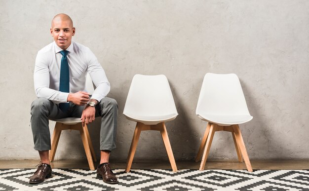 Porträt eines überzeugten glücklichen jungen Geschäftsmannes, der auf Stuhl vor grauer Wand sitzt