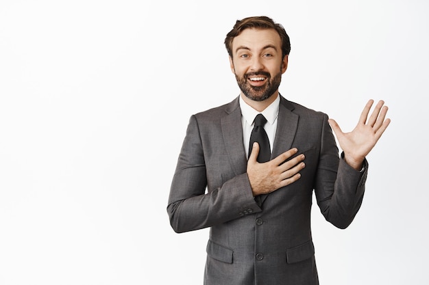 Porträt eines Unternehmensmannes, der die Handfläche hebt, die Hand aufs Herz legt und aufrichtig lächelnd ein Versprechen macht, sich im grauen Anzug vor weißem Hintergrund vorzustellen